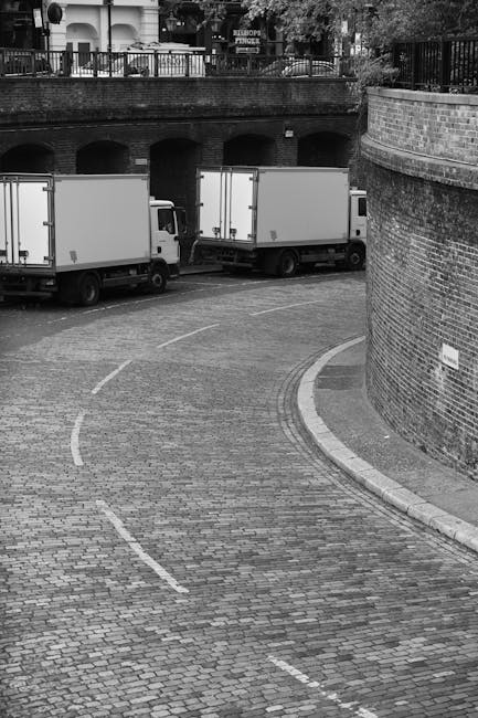 Black and white photograph showing three large white delivery vans parked on a cobbled street near a brick wall and an archway, with an elevated area featuring a metal railing and trees in the background. The vans are positioned parallel to each other, with the rear of the vans facing the curved street, which is marked with dashed white lines indicating traffic lanes. The surrounding environment includes a curved brick wall on the right, and the scene appears to capture a loading or unloading area for house removals or furniture transport, likely part of a home relocation process facilitated by Man with Van Catford. The image emphasizes the movement and logistic aspect of furniture and belongings being prepared for transport, with the vans ready for loading or departure.