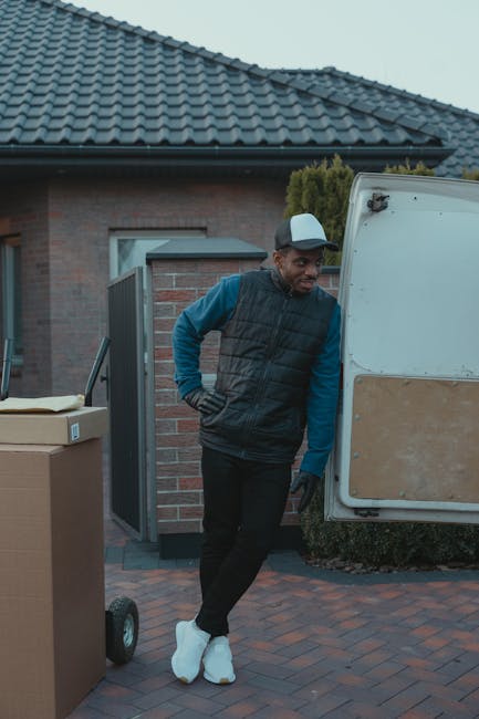 A man wearing a black puffer vest, blue long-sleeve shirt, black trousers, white sneakers, and a grey cap leans against the open rear side of a white moving van parked outside a suburban house with a grey tiled roof and brick exterior. The man appears to be waiting or taking a break during the home relocation process managed by Man with Van Catford, with several cardboard boxes stacked nearby, some on a small hand trolley. The scene is set on a paved driveway adjacent to a garden with bushes, indicating an organized residential move involving furniture transport and loading activities. The natural daylight and outdoor environment suggest the loading process is ongoing or nearing completion as part of packing and moving procedures.