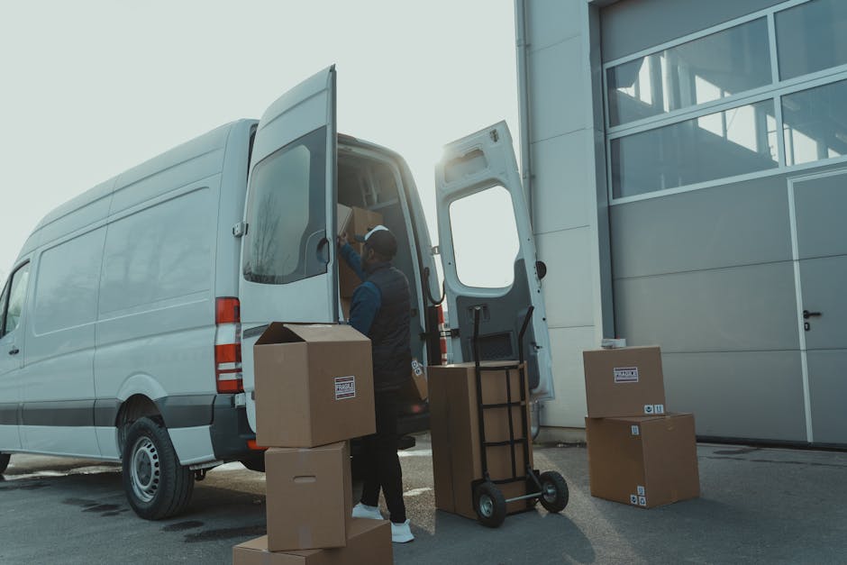 A professional mover from Man with Van Catford is loading cardboard boxes into the rear of a large white van parked outside a modern building with glass windows and a metal facade. The boxes are made of brown corrugated cardboard, some sealed with packing tape, and are arranged on the pavement and on a small hand truck or dolly with black handles, positioned next to the open van door. The mover, dressed in dark clothing and a cap, is inside the van, organizing or placing items inside. The scene depicts the loading process involved in home relocation or furniture transport, with natural daylight illuminating the area and equipment such as blankets or straps potentially visible for securing goods during transit. This process reflects careful packing and loading for a safe and efficient move, characteristic of [COMPANY_NAME]’s removals services supporting reliable house removals and logistics.