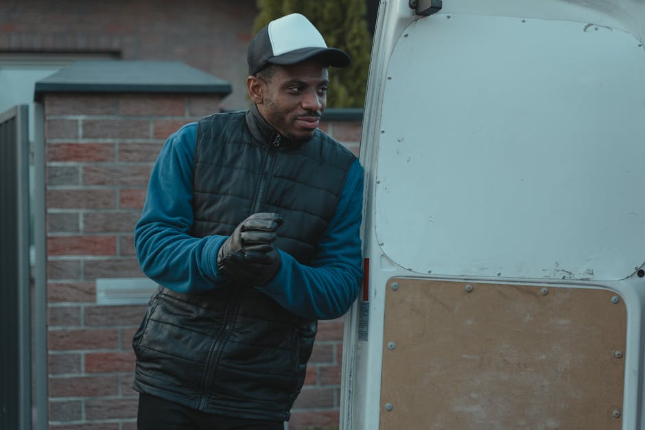 A man wearing a black puffer vest, blue long-sleeve shirt, black trousers, white sneakers, and a grey cap leans against the open rear side of a white moving van parked outside a suburban house with a grey tiled roof and brick exterior. The man appears to be waiting or taking a break during the home relocation process managed by Man with Van Catford, with several cardboard boxes stacked nearby, some on a small hand trolley. The scene is set on a paved driveway adjacent to a garden with bushes, indicating an organized residential move involving furniture transport and loading activities. The natural daylight and outdoor environment suggest the loading process is ongoing or nearing completion as part of packing and moving procedures.